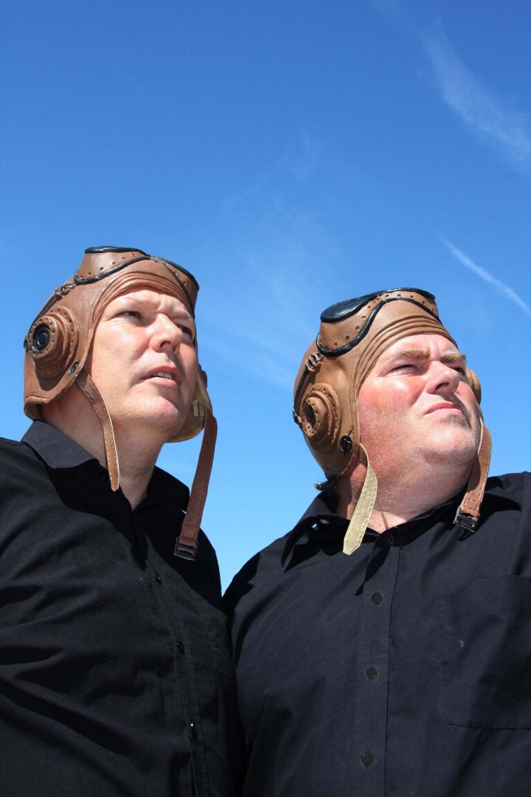 two actors wearing old flying helmets look to the sky