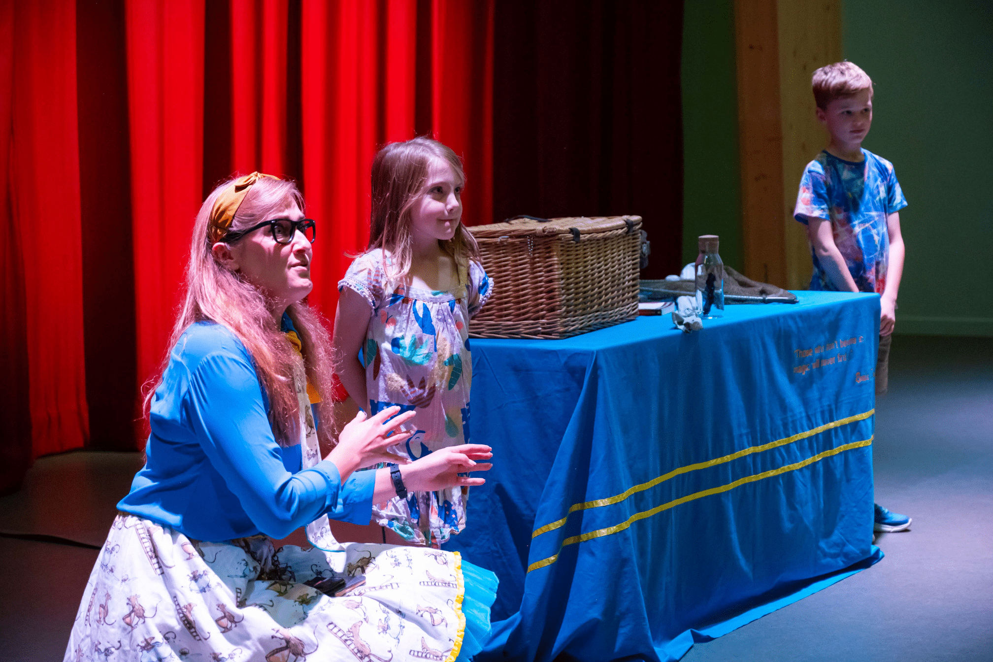 Two children are on stage with a blue table between them. They are with the female actress.