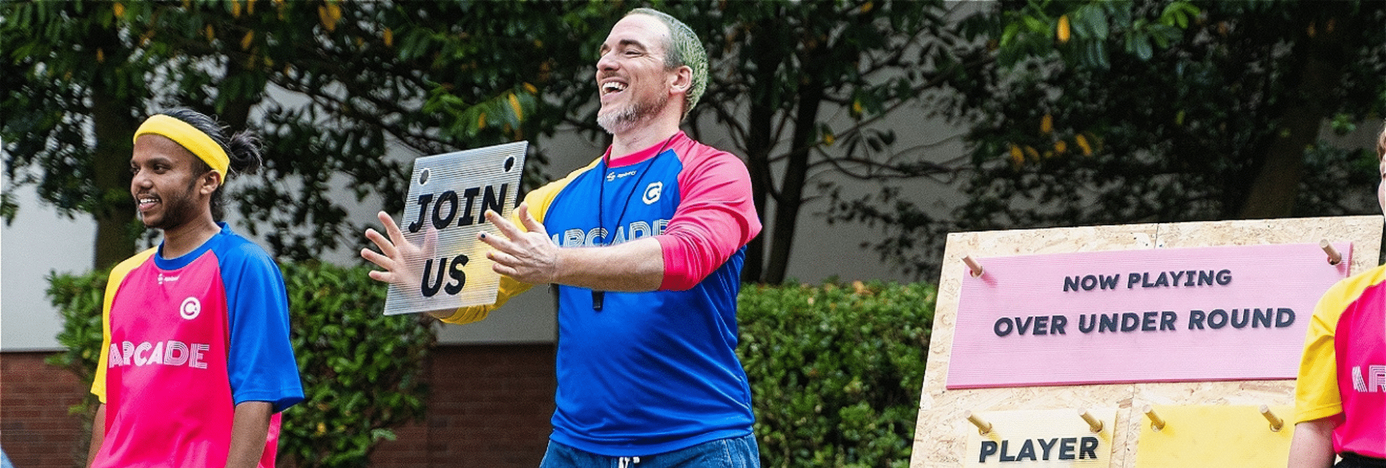 Two men are smiling in brightly coloured tops. One is holding up a sign saying 'join us'. A sign also says 'Now Playing OVER UNDER ROUND' Player 1.