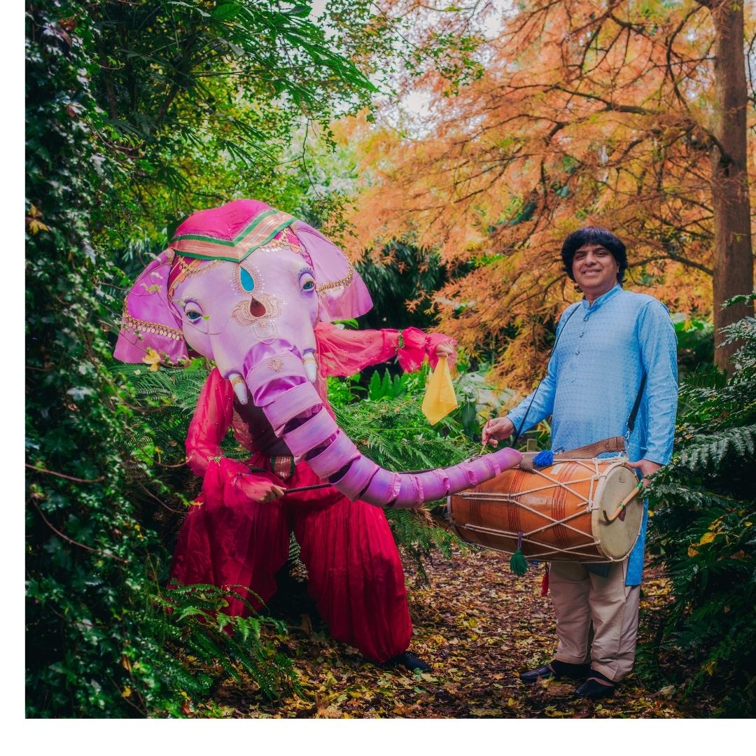 A drummer stands in a wood next to another performer wearing a colourful elephant puppet head.