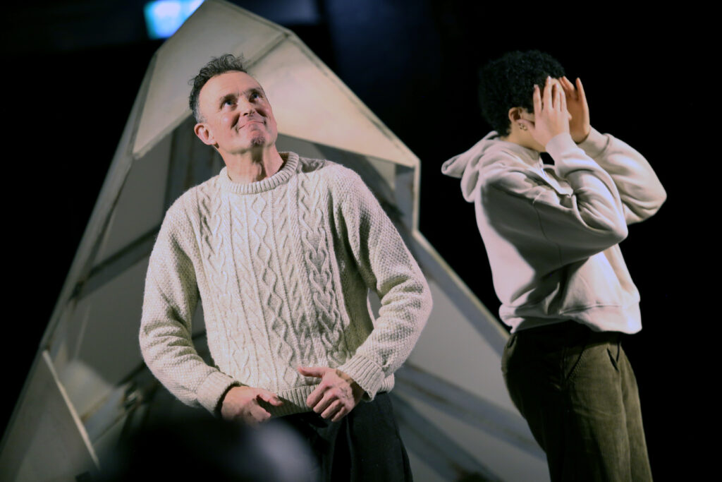 Two actors on stage. One is smiling, the other is covering their face