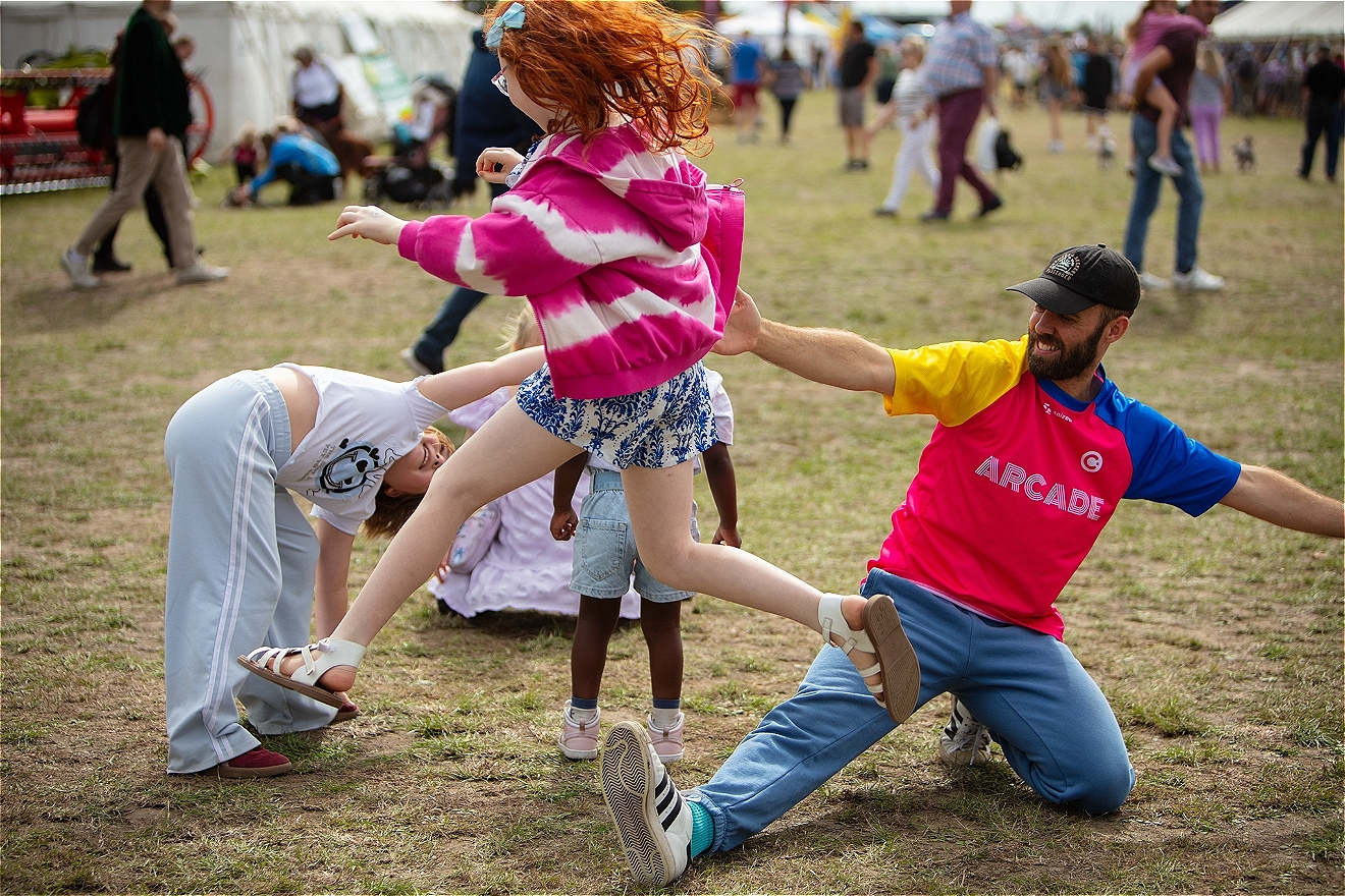 A performer crouches down as a child jumps over them. They are in a field with a crowd behind them