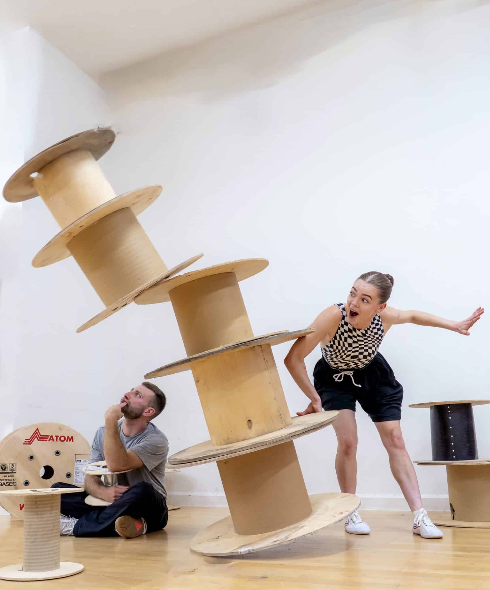 Two performers duck under large wooden reels
