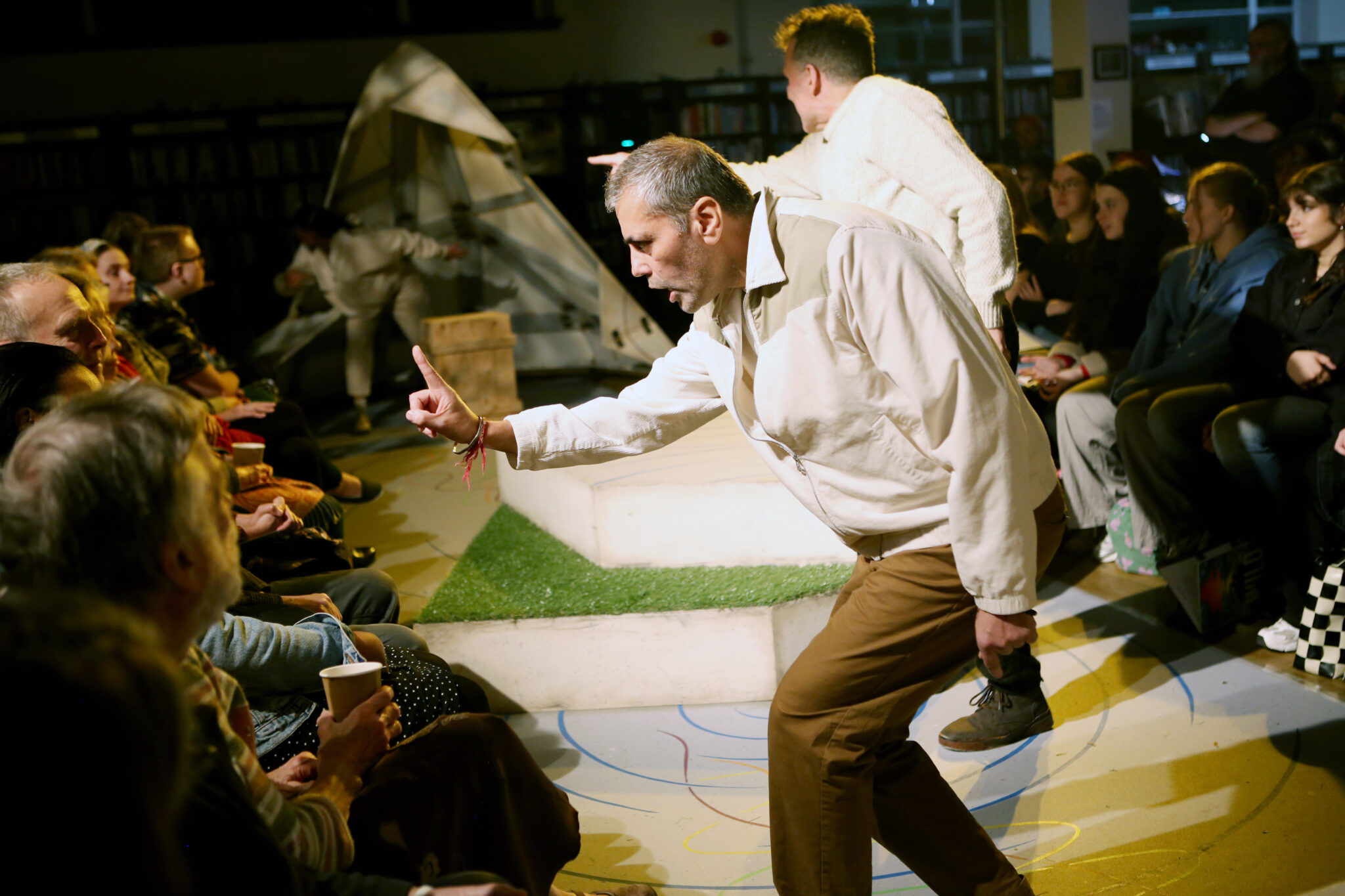 Performer wearing a cream shirt and brown trousers speaks to the audience pointing his finger